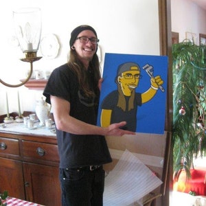 May include: A person holding a blue and yellow painting of a cartoon character wearing a blue hat and holding a wrench. The character is on a blue background. The person is wearing a black shirt, black trousers, and a black hat.