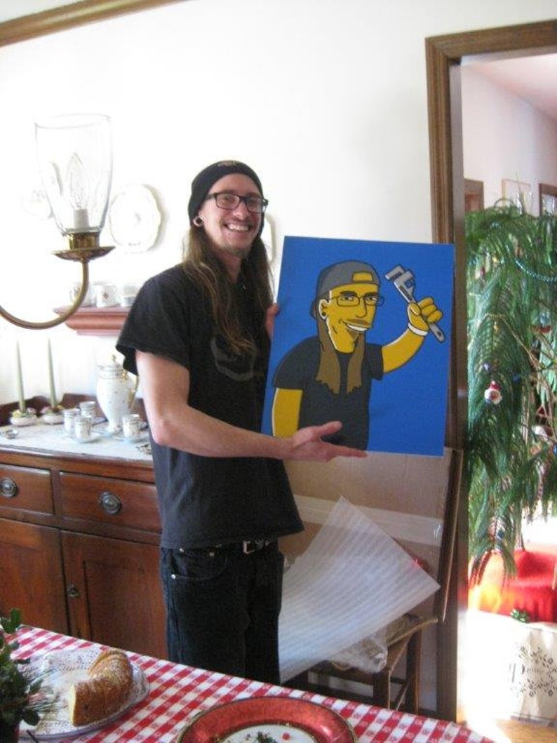 May include: A person holding a blue and yellow painting of a cartoon character wearing a blue hat and holding a wrench. The character is on a blue background. The person is wearing a black shirt, black trousers, and a black hat.