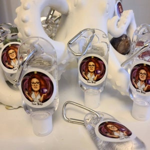 Puede incluir: Cinco botellas de desinfectante de manos de pl&aacute;stico transparente con un mosquet&oacute;n plateado. Cada botella tiene una pegatina redonda con una ilustraci&oacute;n de dibujos animados de una mujer con cabello rojo sobre un fondo morado. El texto "True Node" est&aacute; en cada pegatina.
