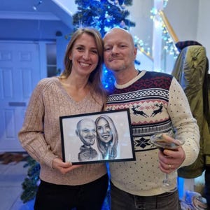 May include: A couple poses for a photo holding a framed black and white caricature drawing of a man and woman. The man is wearing a black shirt and the woman is wearing a white shirt. The couple is standing in front of a Christmas tree with blue lights.