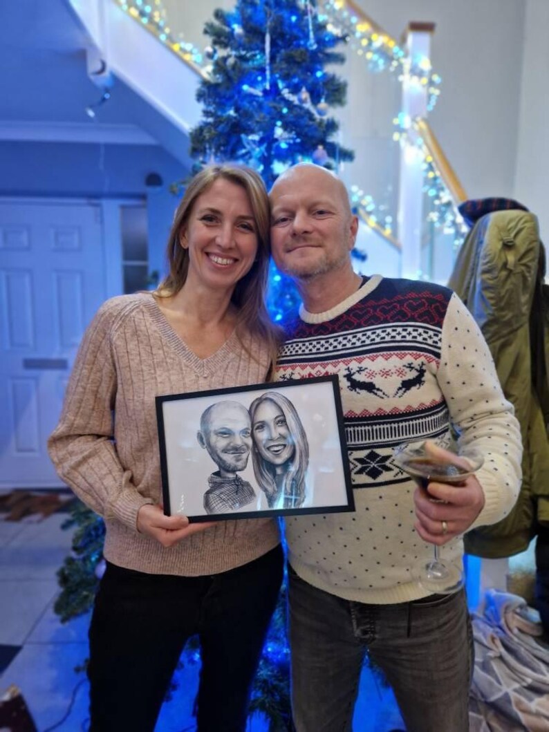 May include: A couple poses for a photo holding a framed black and white caricature drawing of a man and woman. The man is wearing a black shirt and the woman is wearing a white shirt. The couple is standing in front of a Christmas tree with blue lights.