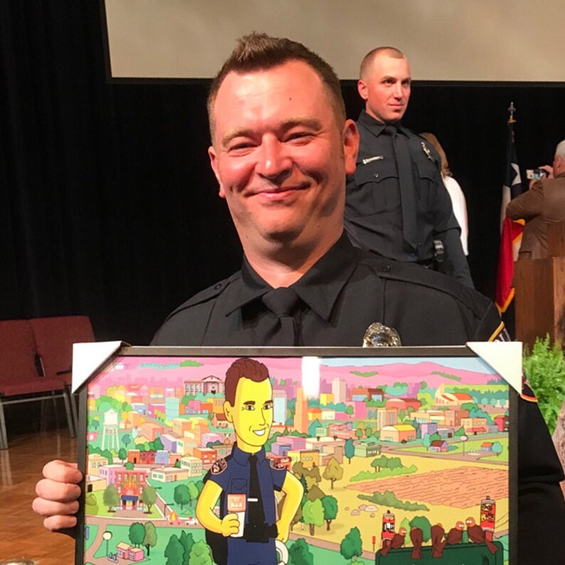 May include: A man in a police uniform smiles while holding a framed cartoon illustration of himself in a police uniform. The cartoon is set against a backdrop of a city skyline with green trees and a pink sky.