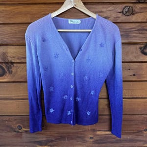 May include: A button-down cardigan in a gradient of purple hues, from light lavender to deep indigo. The long-sleeved sweater features embroidered floral details and is displayed on a wooden hanger against a wood-paneled backdrop. The label reads "Bebeing L".