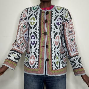 May include: A patterned jacket with a button-up front. The jacket features a mix of colors, including white, pink, green, and purple, with a geometric design. The sleeves are long, and the jacket is worn over a colorful top and blue jeans.