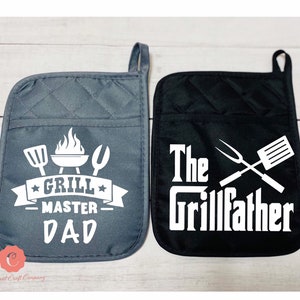 May include: Two quilted oven mitts. One is dark gray with a white graphic of grilling tools and the words "Grill Master Dad". The other is black with the words "The Grillfather" and grilling tools in white.