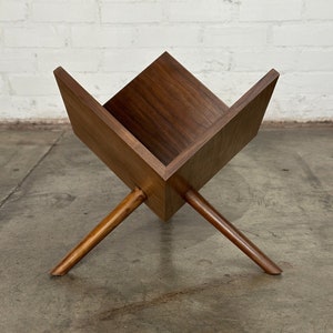May include: A mid-century modern magazine rack made of dark wood. The rack has a geometric design with a square-shaped holder supported by angled legs. The wood grain is visible, and the piece has a warm, natural finish.