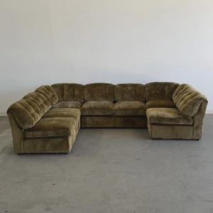 May include: A large, U-shaped sectional sofa in a muted olive green color. The sofa has a low profile and a plush, velvety texture. The design features rounded armrests and a series of individual seat cushions.