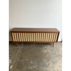 May include: A mid-century modern walnut veneer stereo cabinet with a wood slat front and four legs. The cabinet has a light brown wood finish and a cream-coloured slatted front.