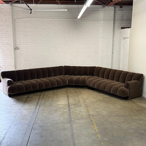 May include: A large brown sectional sofa with tufted upholstery. The sofa is in a warehouse setting with a white brick wall and a concrete floor.