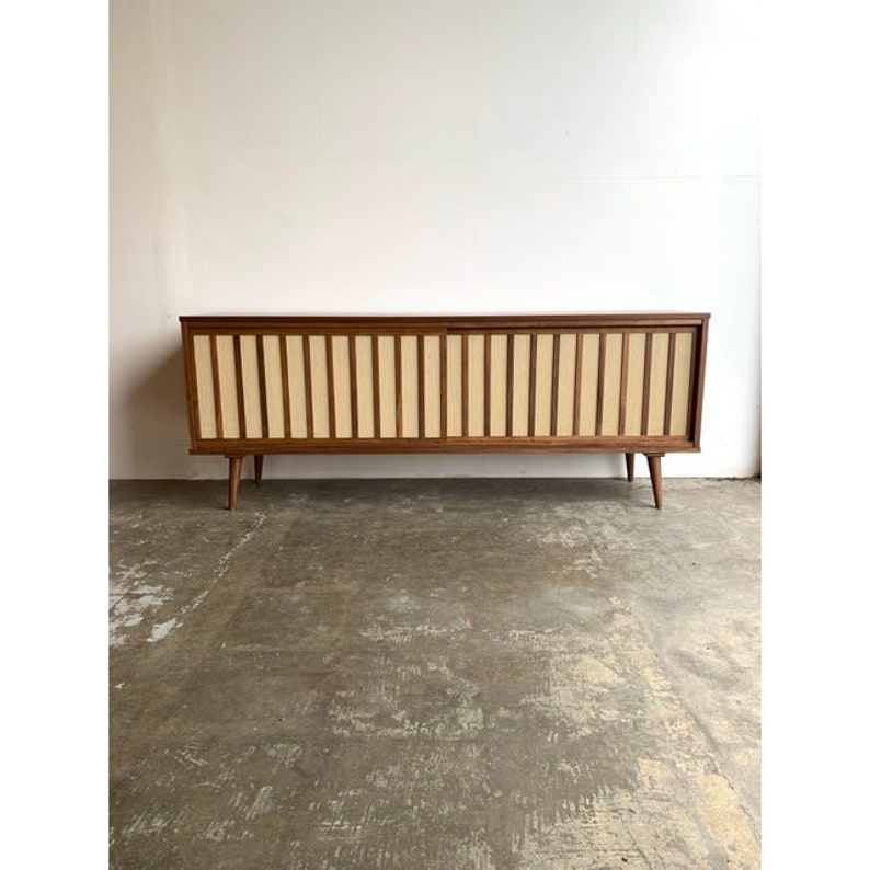 May include: A mid-century modern walnut veneer entertainment console with sliding doors. The doors feature a woven cane panel with a light brown and white pattern. The console has four legs.