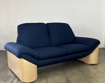 May include: A dark blue two-seater sofa with light wood legs. The sofa has a curved design and is upholstered in a soft fabric.
