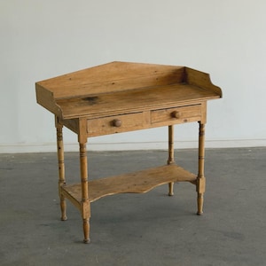 May include: A vintage, light-toned wooden corner table with two small drawers and a lower shelf. The table has a unique, angled design, with turned legs and a distressed finish, showcasing its age and character. The table's dimensions are approximately 30 inches wide, 20 inches deep, and 30 inches high.