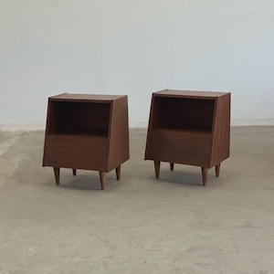 May include: Two mid-century modern nightstands. Each nightstand is made of dark brown wood with a slanted front and a small drawer. The nightstands have three tapered legs and an open shelf.