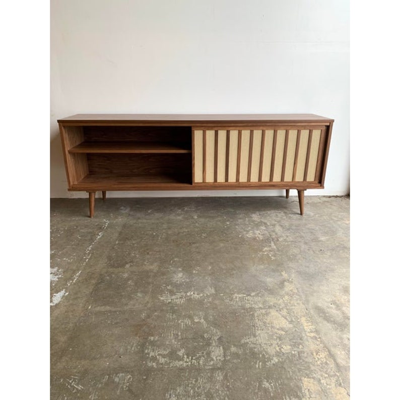 May include: A mid-century modern walnut wood cabinet with a sliding door. The door has a vertical slat design with a light wood veneer. The cabinet has two shelves and sits on four tapered legs.