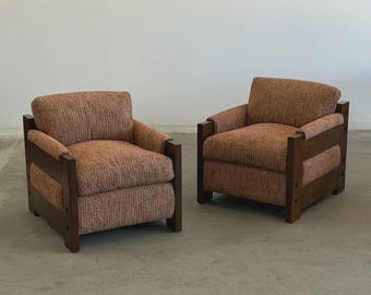 Op de afbeelding: Twee bijpassende fauteuils met een mid-century modern design. De stoelen hebben een donkerbruin houten frame en zijn bekleed met een getextureerde, roodbruine stof. De stoelen zijn vierkant en hebben armleuningen.