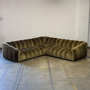 May include: A large, olive green sectional sofa with a unique, ribbed design. The sofa is L-shaped and appears to be made of a soft, velvet-like material. The design features rounded, vertical ridges across the back and arms.