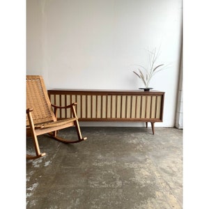 May include: A mid-century modern style wooden cabinet with a light brown wood finish and a light beige fabric front. The cabinet has a series of vertical slats and is sitting on four legs. A rocking chair is in the foreground.