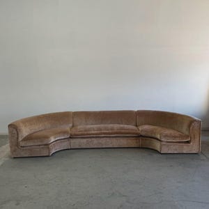 May include: A large, curved sofa in a warm, neutral tone. The sofa has a low profile with a rounded shape, featuring a plush, velvety texture. The design is a sectional, with multiple pieces forming a continuous seating area. The sofa is set against a plain white wall.