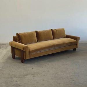 May include: A mid-century modern sofa with a mustard-colored velvet upholstery. The sofa has three back cushions and a rectangular wooden frame. The arms are low and the legs are dark wood. The sofa is a statement piece for any living room.