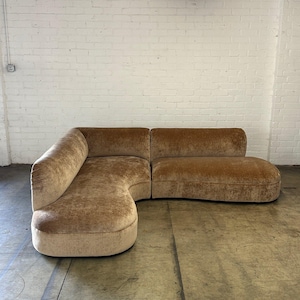 May include: A curved, sectional sofa in a warm, tan color. The sofa has a rounded design with a chaise lounge on one end. The upholstery appears to be a soft, velvety material. The sofa is placed in front of a white brick wall.