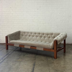 May include: A light brown tufted sofa with a wooden frame and a light gray fabric upholstery. The sofa is sitting on a concrete floor in front of a white brick wall.