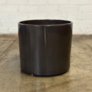 May include: A dark gray ceramic planter with a smooth, rounded surface. The planter is empty and sitting on a concrete floor.