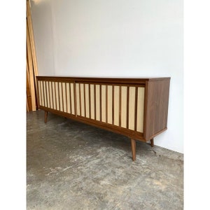 May include: A long, mid-century modern walnut wood sideboard with sliding doors. The doors have a woven fabric panel with vertical wood slats. The sideboard has tapered legs.