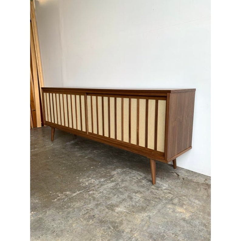 May include: A long, mid-century modern walnut wood sideboard with sliding doors. The doors have a woven fabric panel with vertical wood slats. The sideboard has tapered legs.