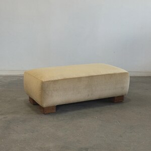May include: A rectangular ottoman with a light beige, velvety fabric. The ottoman sits on four short, rectangular wooden feet. The design is simple and elegant, suitable for a living room or bedroom.