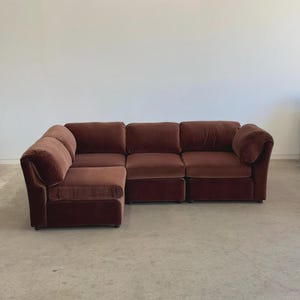 May include: A large, modular sectional sofa in a rich, reddish-brown color. The sofa has a plush, velvet-like texture and a low profile. The design includes an L-shaped configuration with multiple seating sections, perfect for a living room.
