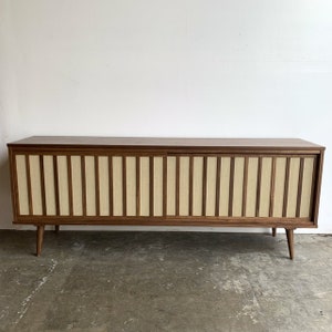 May include: A mid-century modern walnut wood cabinet with sliding doors. The doors are made of a light-coloured woven fabric with a wood frame.