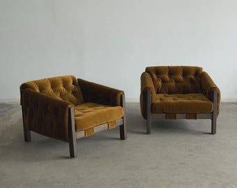 May include: Two matching armchairs with a mid-century modern design. The chairs feature a tufted, mustard-colored velvet upholstery and dark wooden frames. The chairs are set against a plain white wall and a gray concrete floor.