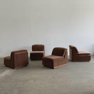 May include: Four brown velvet armchairs. Each chair has a low profile, with a rectangular base and a rounded back. The chairs are arranged in a room with a neutral-colored wall and floor.
