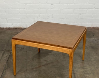 May include: A square wooden coffee table with a light brown wood finish and a simple design. The table has four legs and a flat top.