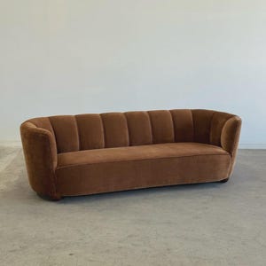 May include: A brown velvet sofa with a curved back and armrests. The sofa has a series of vertical, padded sections across the back and seat. The sofa is set against a plain white wall and sits on a concrete floor.