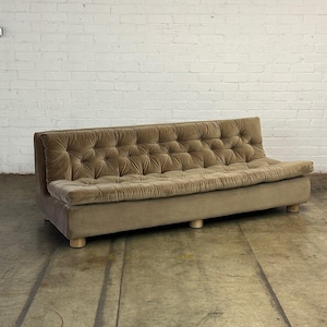 May include: A brown tufted velvet sofa with wooden legs. The sofa is sitting on a concrete floor.
