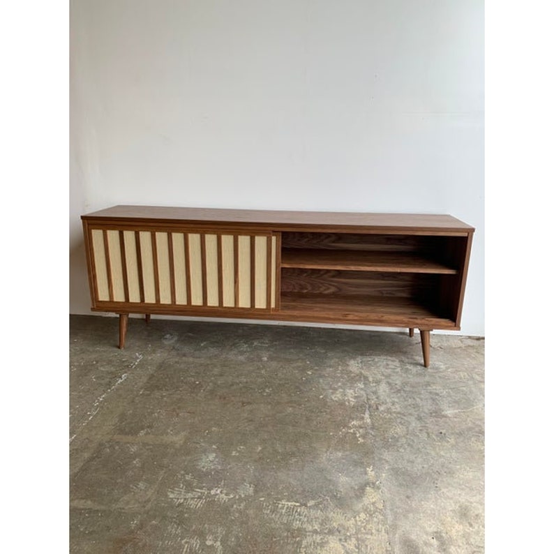 May include: A mid-century modern style walnut wood media console with sliding doors. The doors feature a vertical wood slat design with a light wood inlay. The console has two shelves and sits on four tapered legs.