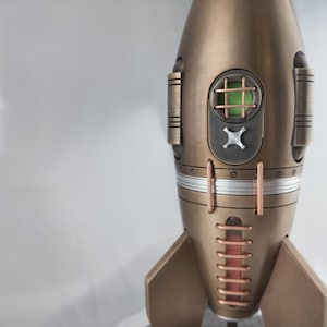 Peut inclure: Une figurine de fusée en bronze avec des accents cuivre et argent. La fusée a une fenêtre verte avec un motif de grille et un X argenté à l'avant.