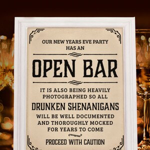 Puede incluir: Un cartel enmarcado en blanco con fondo marrón y texto negro que dice "Nuestra fiesta de Nochevieja tiene un bar abierto. También se está fotografiando mucho, así que todas las travesuras ebrias quedarán bien documentadas y se burlarán durante años. Proceda con precaución."