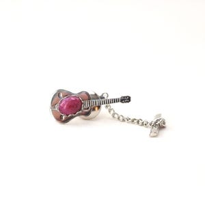 May include: A silver guitar-shaped lapel pin with a large, oval, ruby-colored stone in the center. The pin has a chain and a small, rectangular tag. The guitar body is brown with silver trim.