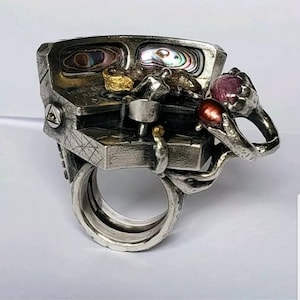 以下が含まれることがあります： アバロンシェルインレイの小さな箱、小さなフィギュア、赤い宝石を特徴とする詳細なデザインのシルバーリング。リングは複数のバンドでできており、テクスチャのある有機的なデザインです。