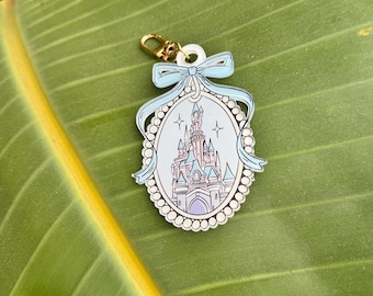 May include: A light blue and white keychain featuring a castle design within an oval frame. The frame is adorned with a light blue bow and a pearl-like border. A gold-colored clasp is attached.