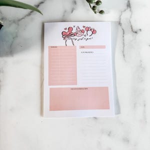 May include: A white notepad with pink sections for to-do lists, date, top priorities, and end-of-day reflection. The notepad features a cartoon illustration with the text "really just a girl!" in black script. The notepad is on a marble surface.