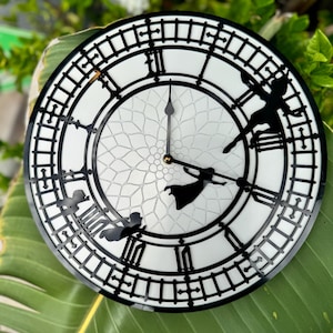May include: A round, white clock with black Roman numerals and a black outer border. The clock hands are black, and the clock face features a decorative pattern. Silhouettes of Peter Pan and Tinkerbell are incorporated into the design. The clock is resting on a large green leaf.