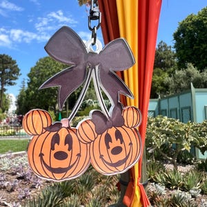 May include: A Halloween-themed keychain featuring two smiling pumpkin-shaped characters with Mickey Mouse ears and a dark gray bow. The keychain is suspended from a metal clasp against a backdrop of a blue sky and autumn decorations.