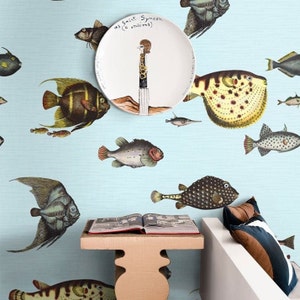 Pu&ograve; includere: Un muro azzurro chiaro con un piatto bianco che presenta un'illustrazione di un personaggio dei cartoni animati in piedi su una roccia. Il piatto &egrave; circondato da un motivo di pesci colorati. Un libro &egrave; su un tavolo di legno davanti a un divano bianco con un cuscino marrone e blu.