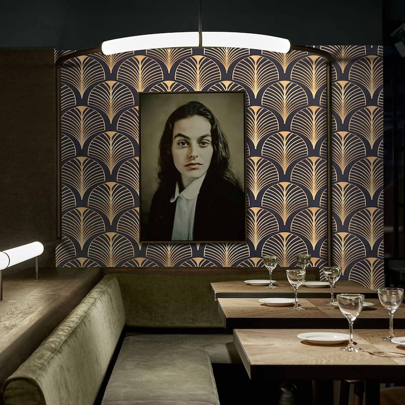 May include: A dark blue and gold geometric wallpaper with a black framed portrait of a person. The wallpaper features a repeating pattern of stylized fans or leaves. The room is decorated with a dark green velvet banquette and wooden tables with glasses and plates.