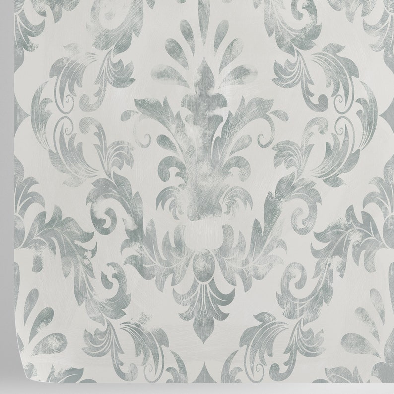 Puede incluir: Primer plano de una muestra de papel pintado con un patr&oacute;n de damasco repetido. El dise&ntilde;o presenta un motivo floral azul gris&aacute;ceo claro sobre un fondo blanco. El patr&oacute;n tiene un aspecto desgastado y texturizado, con una ligera curvatura en el borde.