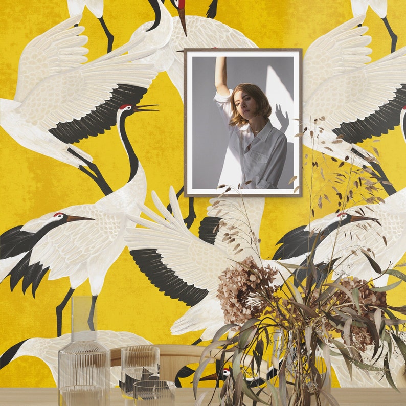 May include: A framed photograph of a person in a white shirt is displayed against a yellow background with a crane pattern. The cranes are white with black wingtips and red accents. A vase of dried flowers and glass containers are in the foreground.