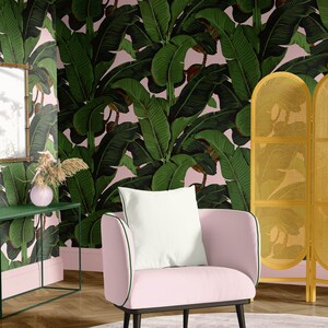 May include: A pink armchair with a white pillow sits in a room with a pink and green banana leaf wallpaper. A gold bamboo screen is in the background.